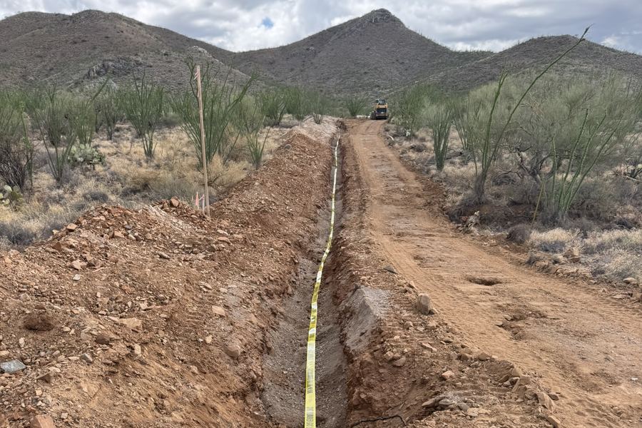 A utility trench cut through Sonoran desert with yellow &ldquo;caution buried gas line below&rdquo; warning tape laid along its length.