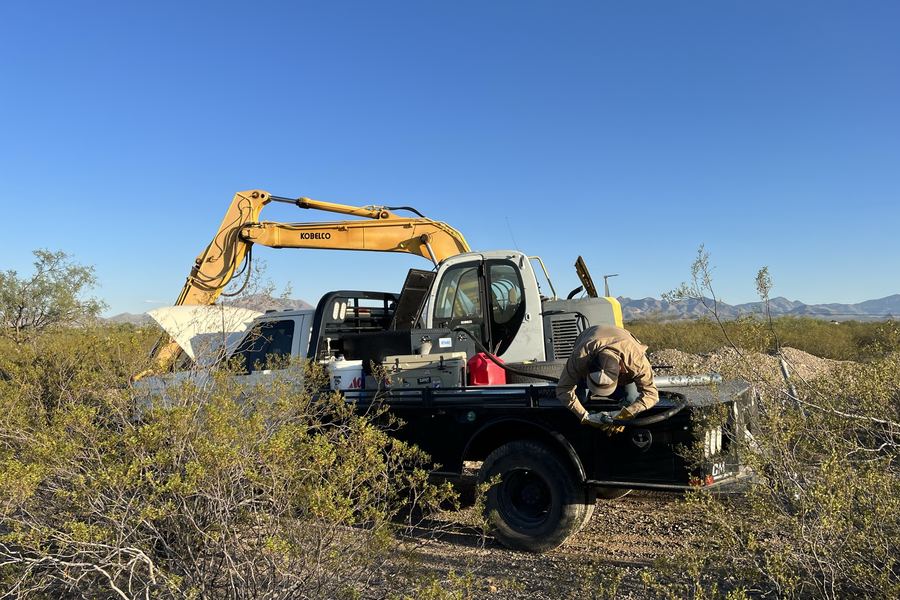 A crew member working at the bed of a flatbed service truck beside a Kobelco excavator on a desert site.